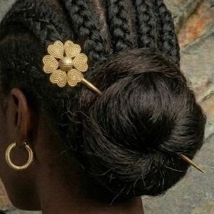 Pode incluir: Penteado feminino com tranças e coque, adornado com um grampo de cabelo dourado em forma de flor. Ela usa brincos de argola dourados e um casaco de veludo verde escuro. O fundo apresenta uma parede de pedra e uma folha de palmeira.
