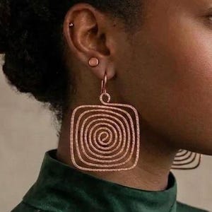 Könnte beinhalten: Ein Paar roségoldfarbene Ohrringe mit einem quadratischen Drahtdesign und einem durchgehenden Spiralmuster. Die Ohrringe hängen an einem einfachen Hakenverschluss und haben eine strukturierte, metallische Oberfläche, die das Licht reflektiert.