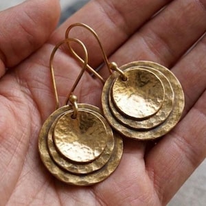May include: A pair of gold-toned drop earrings featuring a layered design of two circular discs. Each disc has a textured, hammered finish. The earrings are suspended from curved wire hooks, creating a dangling effect with a metallic luster.