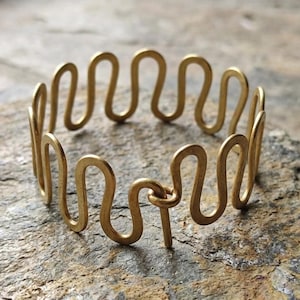 Op de afbeelding: Een goudkleurige armband met een golvend, doorlopend ontwerp. De armband is gemaakt van dun metaaldraad en heeft een unieke, artistieke uitstraling. De armband heeft een diameter van ongeveer 6,35 cm.