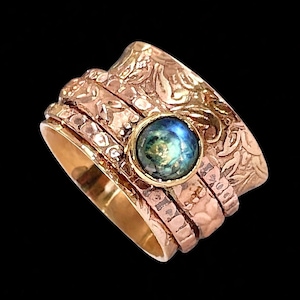 Op de afbeelding: Een goudkleurige ring met een centrale, iriserende, blauwgroene steen. De ring heeft meerdere banden met getextureerde en gegraveerde details, waaronder bloemmotieven en gehamerde afwerkingen. De ring staat tegen een zwarte achtergrond.