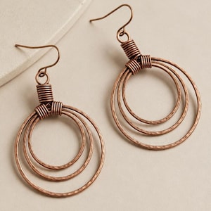 May include: A pair of copper-toned dangle earrings featuring three concentric circles suspended from a wire hook. The metal surface of each hoop has a textured, hammered finish, and the circles are secured together by tightly coiled wire.