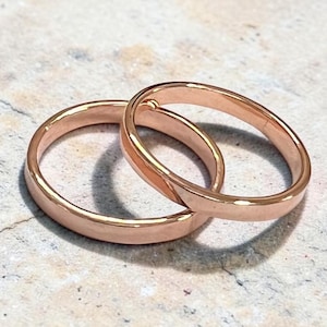 Peut inclure: Deux alliances en or rose, entrelacées, posées sur une surface texturée et claire. Les anneaux sont lisses et brillants, reflétant la lumière. Les bagues sont simples et élégantes, de conception classique.