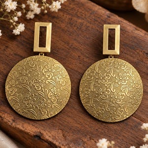 Pode incluir: Um par de brincos dourados com um grande pendente circular que apresenta um intrincado padrão floral em relevo. Cada peça circular está suspensa por uma pequena moldura retangular dourada, criando um design pendente com acabamento mate.