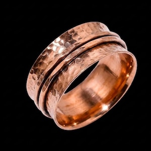 May include: A copper spinner ring with a hammered finish. The ring has a wide band with three rotating sections. The metal has a warm, reddish-brown hue. The design is simple but striking, suitable for everyday wear.