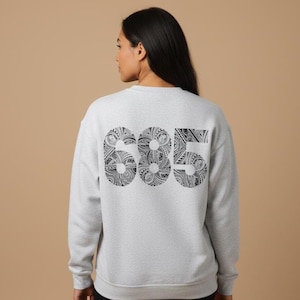 May include: A light gray sweatshirt with the number "685" printed on the back in black. The numbers are filled with a detailed, tribal-inspired pattern. The sweatshirt is a casual, comfortable style.