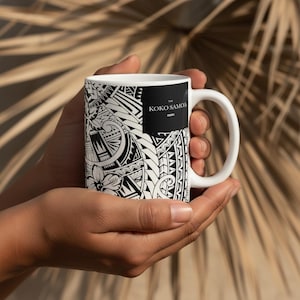 May include: White ceramic mug featuring a black and white tribal pattern and the text "The Koko Samoa". The mug has a curved handle and is held in two hands.