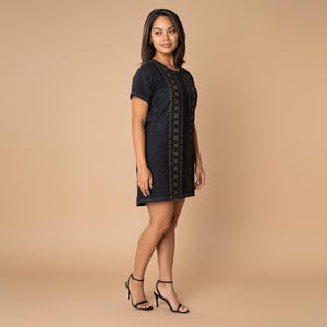 Pode incluir: Um vestido preto de manga curta com um painel vertical de intrincados bordados geométricos dourados ao centro. O vestido tem um decote redondo e uma bainha reta que cai acima do joelho, combinado com sandálias pretas de salto alto.