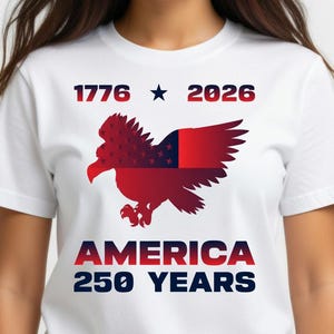 Pode incluir: T-shirt branco com um design patriótico. O design inclui o texto "1776 ★ 2026" acima de um gráfico de águia vermelho, branco e azul. Abaixo da águia, o texto diz "AMERICA 250 YEARS".