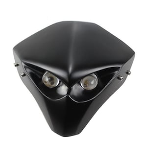 May include: A black motorcycle headlight assembly with two round, clear lenses. The housing has a matte black finish and a pointed, angular design. Small silver fasteners are visible.