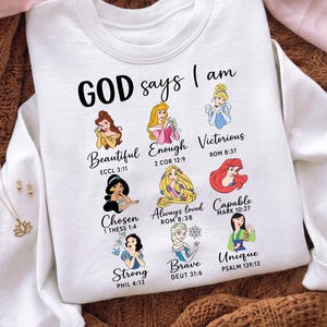 May include: A white crewneck sweatshirt featuring the text "GOD says I am" above nine illustrated female characters. Each character is paired with a descriptive word and a Bible verse reference, including "Beautiful," "Enough," "Victorious," "Chosen," "Always loved," "Capable," "Strong," "Brave," and "Unique."