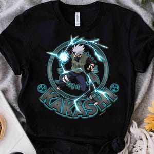 May include: A black short-sleeve t-shirt featuring a graphic of the anime character Kakashi Hatake surrounded by blue lightning bolts. The design includes a circular emblem, the name KAKASHI in bold letters, and small shuriken symbols.