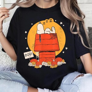 May include: A black t-shirt featuring a graphic of Snoopy sleeping on a red doghouse against a large yellow moon. The design includes orange pumpkins, spiderwebs, and a sign that reads BOO! with the letter Z repeated above the doghouse.