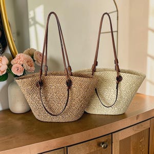 以下が含まれることがあります： 茶色の革の持ち手が付いた、2つの織り込まれたストローのトートバッグ。1つのバッグは薄いタン色で、もう1つは自然なストロー色です。バッグは木製の表面に置かれています。