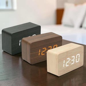 May include: Three rectangular wooden digital clocks in black, brown, and light wood tones. The clocks display the time in a bright digital font. The clocks are on a dark wooden surface.