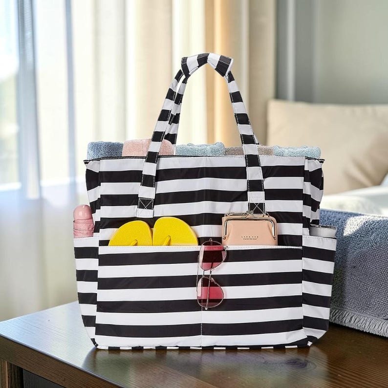 Może przedstawiać: Czarno-biała torba typu tote w paski z wieloma kieszeniami. Torba zawiera ż&oacute;łte klapki, r&oacute;żowy portfel, okulary przeciwsłoneczne i zwinięte ręczniki. Torba stoi na drewnianej powierzchni.