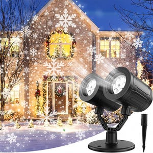 May include: A black dual-lens LED light projector with a circular base and a separate ground stake. The device projects white snowflake patterns onto the exterior of a brick house, which is decorated with festive wreaths and warm interior lighting.