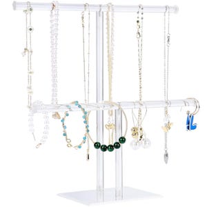 May include: Clear acrylic jewelry display stand with multiple bars for hanging necklaces, bracelets, and earrings. The stand is rectangular at the base and has a vertical support with horizontal bars. Various necklaces and bracelets are displayed.
