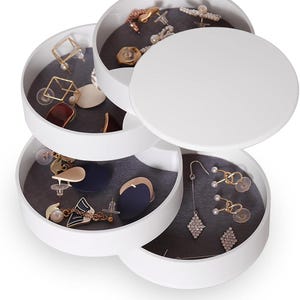 May include: A white, multi-tiered, rotating jewelry organizer with four circular compartments. Each tray features a dark gray velvet lining and contains various earrings, including gold-toned geometric shapes, pearl studs, and dangling crystal pieces.