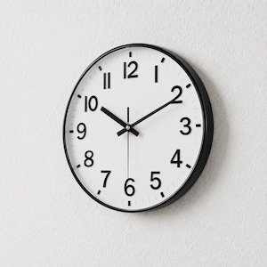Puede incluir: Un reloj de pared redondo con esfera blanca y números y manecillas negras. El reloj tiene un marco negro y está montado en una pared blanca. La hora que se muestra es aproximadamente las 10:10.