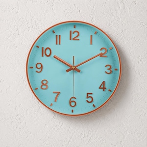 Op de afbeelding: Een ronde wandklok met een turquoise wijzerplaat en koperkleurige wijzers en cijfers. De klok heeft een koperkleurige rand en staat tegen een witte, getextureerde muur. De wijzers geven de tijd aan.