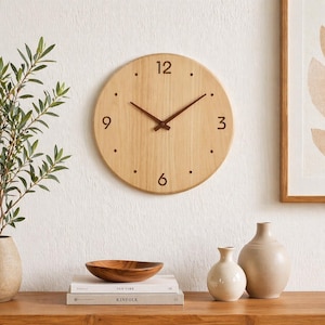 Peut inclure: Une horloge murale ronde en bois clair avec des aiguilles marron et des points comme marqueurs d'heures. L'horloge est fixée sur un mur blanc. En dessous, une table en bois avec des livres, un bol en bois et deux vases en céramique.