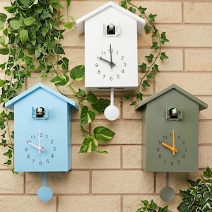 May include: Three wooden cuckoo clocks in white, blue, and green, each with a house-shaped design. The clocks have white clock faces with black numbers and hands, and a pendulum. A small bird is visible in the opening.