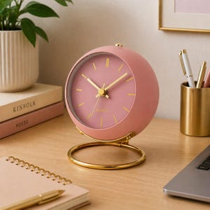 May include: A pink quartz analog clock with gold-colored hands and a gold base. The clock has a round face with gold markers and the word "QUARTZ" printed at the bottom. The clock is sitting on a wooden desk.