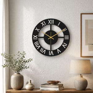 Op de afbeelding: Een zwarte wandklok met gouden wijzers en Romeinse cijfers. De klok is rond met een minimalistisch ontwerp, gemonteerd op een witte, getextureerde muur. De wijzers van de klok geven de tijd aan. Het is een decoratief item.