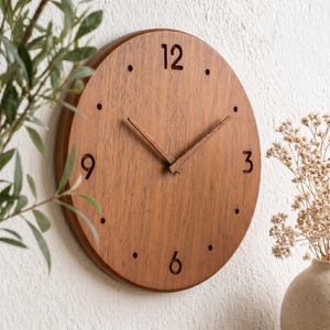 Op de afbeelding: Een ronde houten wandklok met een natuurlijke houtnerf. Zwarte cijfers markeren de uren 12, 3, 6 en 9, met stippen voor de andere uren. De wijzers van de klok zijn ook van hout.