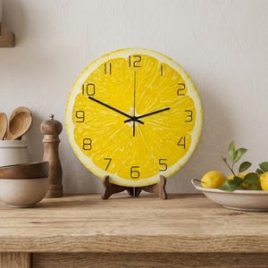 Op de afbeelding: Een ronde wandklok met een heldergeel citroen schijf ontwerp. De klok heeft zwarte wijzers en cijfers en wordt op een houten standaard getoond. Een decoratief keukenartikel.