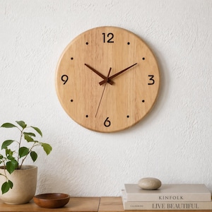 Peut inclure: Une horloge murale ronde en bois clair. L'horloge présente des chiffres noirs aux positions 12, 3, 6 et 9 heures, avec des points marquant les autres heures. Les aiguilles sont marron foncé. L'horloge mesure environ 30 cm de diamètre.