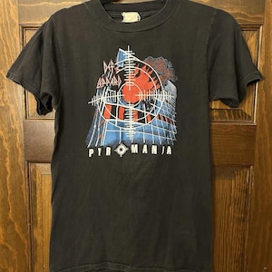 Könnte beinhalten: Schwarzes T-Shirt mit einem grafischen Design mit dem Text "PYROMANIA". Das Design umfasst ein rotes Ziel, ein Gebäude und den Bandnamen "DEF LEPPARD". Das T-Shirt ist kurzärmlig.