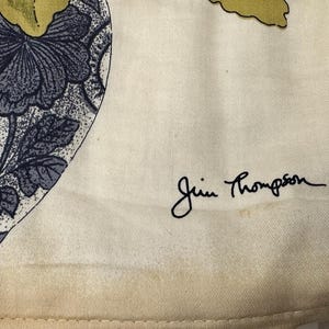 Pode incluir: Tecido creme com um padr&atilde;o floral em tons de azul e cinza. O design apresenta grandes flores e folhas. A assinatura "Jim Thompson" est&aacute; escrita em escrita preta. O tecido tem uma apar&ecirc;ncia ligeiramente envelhecida.