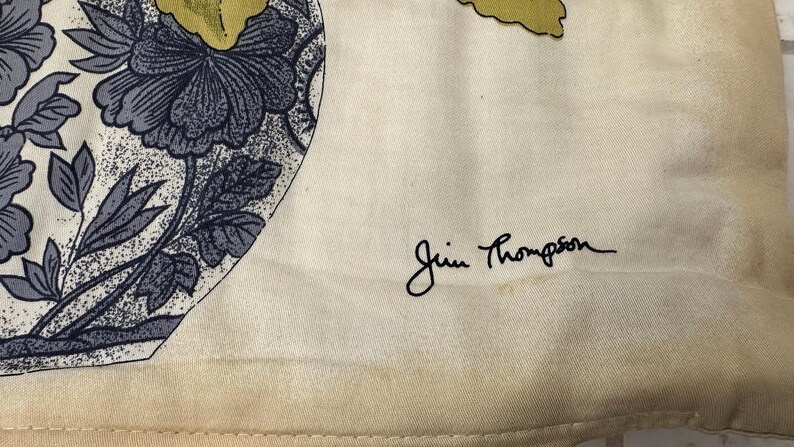 Pode incluir: Tecido creme com um padr&atilde;o floral em tons de azul e cinza. O design apresenta grandes flores e folhas. A assinatura "Jim Thompson" est&aacute; escrita em escrita preta. O tecido tem uma apar&ecirc;ncia ligeiramente envelhecida.