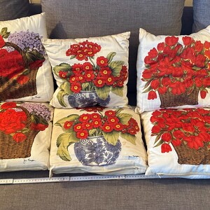 Pode incluir: Seis almofadas decorativas com desenhos florais. As almofadas apresentam flores vermelhas vibrantes em cestos e vasos de vime, sobre um fundo creme. As almofadas est&atilde;o dispostas num sof&aacute; cinzento.