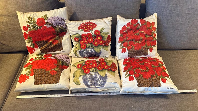 Pode incluir: Seis almofadas decorativas com desenhos florais. As almofadas apresentam flores vermelhas vibrantes em cestos e vasos de vime, sobre um fundo creme. As almofadas est&atilde;o dispostas num sof&aacute; cinzento.