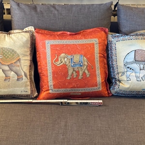 Puede incluir: Tres cojines decorativos con dise&ntilde;os de elefantes. Los cojines presentan ilustraciones detalladas de elefantes en varias poses, con patrones ornamentados y colores vibrantes. Los cojines son cuadrados y descansan sobre un sof&aacute; gris.