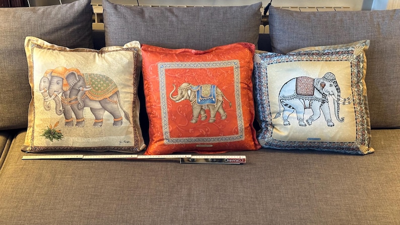 Puede incluir: Tres cojines decorativos con dise&ntilde;os de elefantes. Los cojines presentan ilustraciones detalladas de elefantes en varias poses, con patrones ornamentados y colores vibrantes. Los cojines son cuadrados y descansan sobre un sof&aacute; gris.