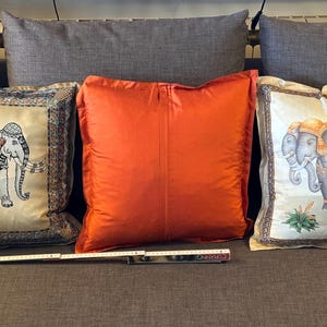 Puede incluir: Tres cojines decorativos sobre un sof&aacute; gris. Dos cojines presentan dise&ntilde;os de elefantes en beige y dorado, mientras que el coj&iacute;n central es de color naranja liso. Los cojines son rectangulares y miden unos 45 cm cuadrados.