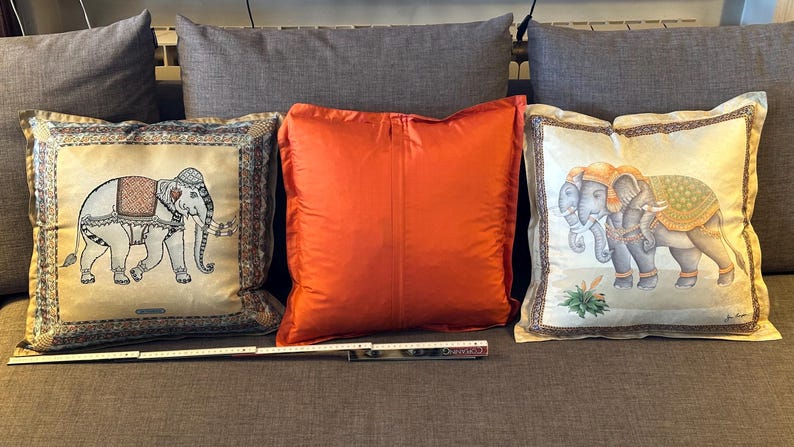 Puede incluir: Tres cojines decorativos sobre un sof&aacute; gris. Dos cojines presentan dise&ntilde;os de elefantes en beige y dorado, mientras que el coj&iacute;n central es de color naranja liso. Los cojines son rectangulares y miden unos 45 cm cuadrados.