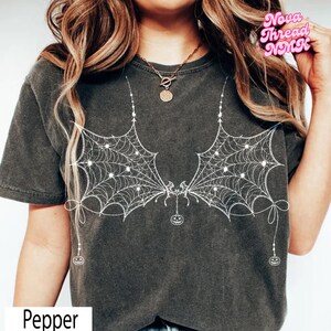 Könnte beinhalten: Ein anthrazitfarbenes Kurzarm-T-Shirt mit einem weißen Grafikdruck von zwei symmetrischen Fledermausflügeln, die aus Spinnenweben bestehen. Das Design enthält kleine hängende Kürbisse an den Flügeln sowie dekorative Schleifen an den Seiten.