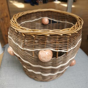 Könnte beinhalten: Ein runder geflochtener Korb aus braunen Weidenruten mit eingearbeiteten horizontalen Bändern aus hellerem, cremefarbenem Material. Der Korb ist mit mehreren glatten, polierten, pfirsichfarbenen Steinkugeln verziert, die an der Außenseite befestigt sind.