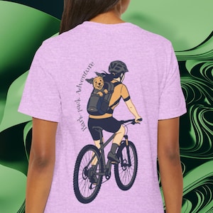 May include: A light purple t-shirt featuring a graphic of a person riding a mountain bike with a dog in a backpack. The text "Bark pack Adventure" is printed vertically on the left side of the shirt.