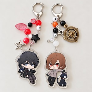 Può includere: Due portachiavi in acrilico con personaggi in stile anime. Il ciondolo a sinistra mostra un personaggio dai capelli neri con un gatto, decorato con perline rosse, una foglia rosa e una stella nera. Quello a destra presenta un personaggio dai capelli castani con un gatto, un ciondolo a forma di bussola in bronzo e perline arancioni, nere e bianche.
