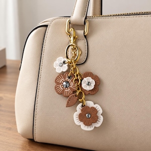 May include: A beige handbag featuring a gold-toned metal keychain attachment. The keychain consists of a gold chain adorned with multiple layered flower-shaped charms in white and brown, each featuring a small central stud detail.
