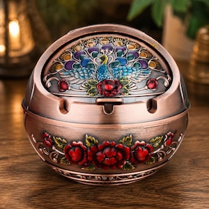 May include: A round, copper-toned metal ashtray featuring a rotating lid with an embossed peacock design in blue, green, and purple enamel. The base is decorated with raised, hand-painted red floral motifs and green leaves in a metallic finish.