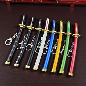 Può includere: Una collezione di otto portachiavi a forma di spada con guardia e pomolo color oro. I foderi sono disponibili in vari colori, tra cui nero con motivi bianchi, bianco, blu, giallo, verde e rosso, ciascuno dotato di un anello in metallo.