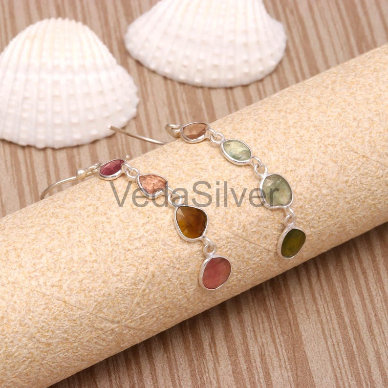 The same multi-tourmaline earrings resting on a textured display roll next to two white seashells.