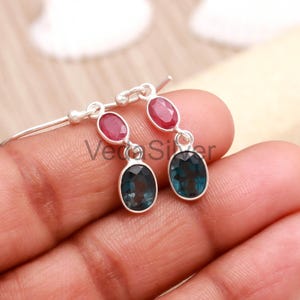 May include: A pair of silver earrings featuring a dangling design. Each earring has an oval-shaped, deep blue gemstone suspended below a smaller, oval-shaped, red gemstone. The earrings are held by a person's hand.